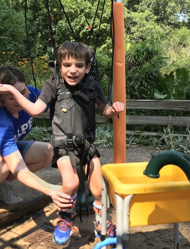 Child in a harness system with an adult who is helping place the child's right foot on the ground. 