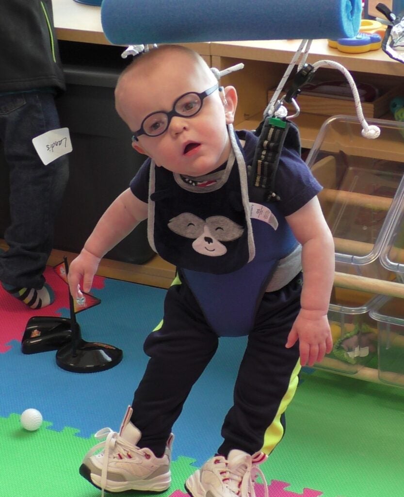 Toddler in a harness standing on a play mat.